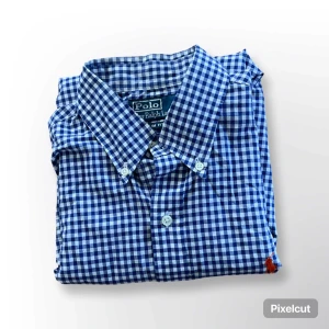 Blårutig skjorta från Polo Ralph Lauren - Klassisk blårutig skjorta från Polo Ralph Lauren med vit knappstängning och krage. Skjortan har lång ärm och är tillverkad i ett mjukt bomullstyg. Perfekt för en stilren och avslappnad look.