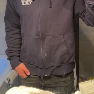 Mörkblå hoodie med dragkedja från Norrlands Guld - Mörkblå hoodie med dragkedja och huva från Norrlands Guld. Tröjan har fickor framtill och en broderad logga på bröstet. Perfekt för en avslappnad stil och enkel att matcha med jeans eller joggers. Pris kan diskuteras
