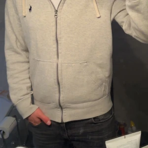 Ljusgrå hoodie från Polo Ralph Lauren - Säljer en stilren ljusgrå hoodie från Polo Ralph Lauren med dragkedja och huva. Tröjan har två fickor framtill och en liten broderad logga på bröstet. Perfekt för en avslappnad och snygg look. Köpt för 1800 men knappt använd. Pris är ej hugget i sten tveka inte på o fråga peace out