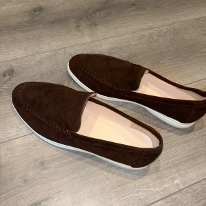 Bruna mocka loafers med vit sula - Säljer ett par stilrena bruna loafers i mocka med vit platt sula. Skorna har en klassisk design med rund tå och diskreta sömmar längs ovandelen. Perfekta för en avslappnad men snygg look. Inte använts nån gång!