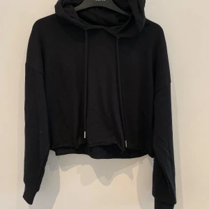 Svart croppad hoodie - Säljer en svart croppad hoodie med huva och långa ärmar. Tröjan har dragsko i halsen och är tillverkad i mjukt material. Perfekt för en avslappnad stil.