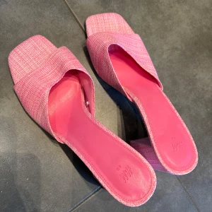 Rosa sandaler med klack från H&M - Säljer ett par trendiga rosa sandaler från H&M med bred rem och fyrkantig tå. Skorna har en hög blockklack och är tillverkade i ett vävt syntetmaterial med rosa innersula. Perfekta för att lyfta din outfit!