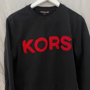 Svart sweatshirt från Michael Kors med röd logga - Svart sweatshirt från Michael Kors med stora röda bokstäver 'KORS' broderat på bröstet. Tröjan har rund halsringning och långa ärmar. Perfekt för en stilren och trendig look.