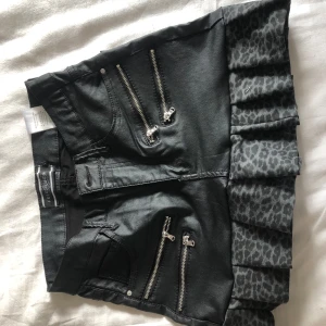 Svart kjol med leopardvolang och dragkedjor - Cool svart kjol från Bershka med flera silvriga dragkedjor framtill och volangkant i grått leopardmönster. Kjolen har jeanslook och två bakfickor. Perfekt för dig som vill sticka ut med en edgy stil.