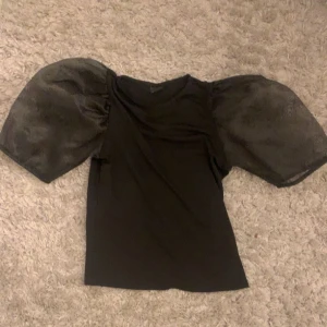 Svart topp med puffärmar - Säljer en svart topp med stora puffärmar i transparent material. Toppen har rund halsringning och kort ärm med volym, vilket ger en trendig och cool look.