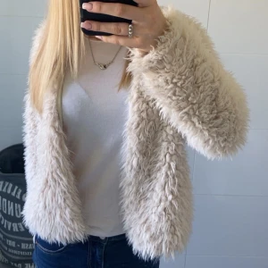 Beige pälsjacka - Säljer en fluffig och mjuk beige pälsjacka med lång ärm. Jackan har en öppen front utan synliga knappar eller dragkedja och ger en trendig look. Perfekt för dig som vill sticka ut med en mysig och stilren jacka.