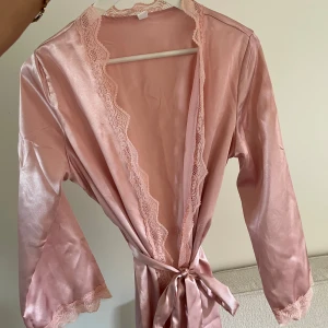 Rosa satin morgonrock med spetsdetaljer - Säljer en ljusrosa morgonrock i satin med fina spetsdetaljer längs kanterna och ärmsluten. Rocken har trekvartsärm och knyts i midjan med ett matchande bälte. Perfekt för en lyxig känsla hemma.