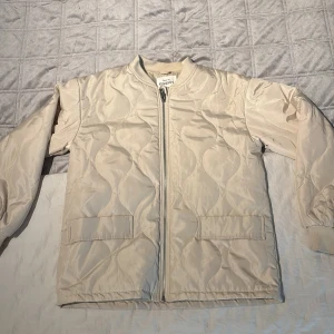 Beige quiltad jacka från KappAhl - Beige quiltad jacka från KappAhl – strl 146  Säljer en helt ny, oanvänd quiltad jacka från KappAhl i storlek 146. Jackan har långa ärmar, dragkedja framtill, två ytterfickor och en liten innerficka – perfekt för att förvara nycklar eller småsaker. Stilren design med ett diskret quiltat mönster, perfekt för övergångsperioder som vår och höst. Jackan är i helt nyskick, endast prislappen som saknas.