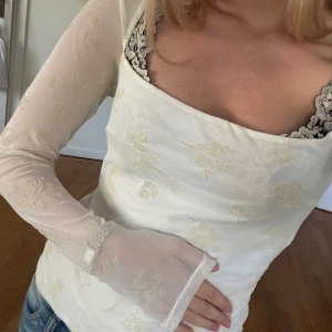 Vit långärmad blus med blommigt mönster - Säljer en vit långärmad blus med diskret blommigt mönster och transparenta ärmar. Toppen har en fyrkantig urringning och är i ett tunt, lätt material. Perfekt att styla med jeans för en trendig look.