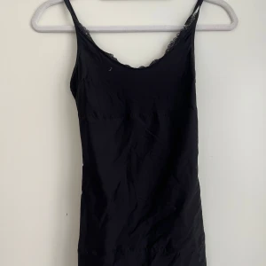Svart linnetopp med spetskant - Säljer en svart linnetopp med tunna axelband och fin spetsdetalj längs urringningen. Perfekt att styla till olika outfits och enkel att bära under andra plagg. Den formar kroppen väldigt fint!