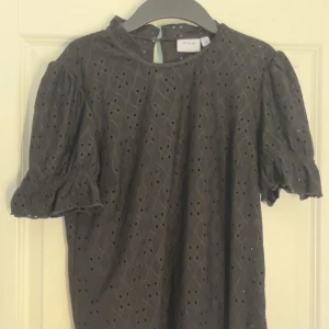 Svart blus med detaljer - Säljer en svart blus från Vila med broderade hålmönster och puffärmar. Blusen har rund halsringning, korta ärmar med resår och en liten knapp i i nacken!