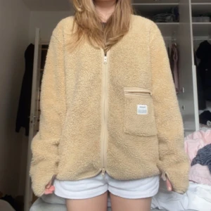 Beige teddyjacka från Resteröds - Mysig beige teddyjacka från Resteröds med dragkedja framtill och en stor ficka med dragkedja på bröstet. Jackan har en oversized passform och är perfekt för kyligare dagar. Fleece-liknande material som känns mjukt och fluffigt.