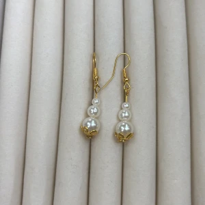Guldiga örhängen med pärlor - Säljer ett par eleganta örhängen med tre vita pärlor i olika storlekar och guldfärgade detaljer. Perfekta för att lyfta din stil med en klassisk touch.