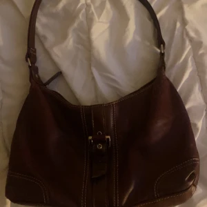 Vinröd vintage axelväska i skinn - Säljer en stilren vinröd axelväska i skinn med dekorativa sömmar och spänndetalj framtill. Väskan har ett huvudfack och en mindre ficka med dragkedja på baksidan. Perfekt för dig som vill ha en klassisk accessoar.