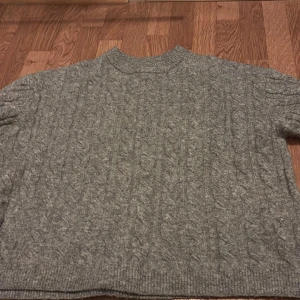 Grå kabelstickad tröja - Mysig grå kabelstickad tröja med rund hals och långa ärmar. Perfekt för kyligare dagar och enkel att matcha med olika outfits. Klassisk design med ribbade muddar vid ärmslut och nederkant.