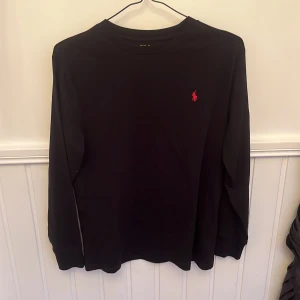 Svart långärmad tröja från Polo Ralph Lauren - Säljer en svart långärmad tröja från Polo Ralph Lauren med den klassiska röda broderade loggan på bröstet. Tröjan har en enkel och stilren design, perfekt för en clean look. Mjuk och skön bomullskvalitet.