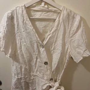 Vit omlottopp i linne från H&M - Säljer en vit omlottopp i linne från H&M med korta ärmar och knyt i midjan. Toppen har v-ringning och knäpps med knappar framtill.