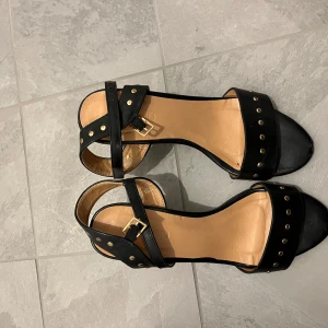 Svarta sandaler med nitar och öppen tå - Säljer ett par svarta sandaler med öppen tå och klack. Skorna har guldfärgade nitar längs remmarna och ett spänne vid ankeln. Perfekta för dig som vill ha en stilren men edgy look.