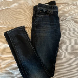 Mörkblå jeans från Jack & Jones - Säljer ett par mörkblå jeans från Jack & Jones med klassisk femficksdesign och diskreta slitningar. Jeansen har normal passform och raka ben, perfekt för en avslappnad stil. Snygga kontrastsömmar och traditionell dragkedja framtill.