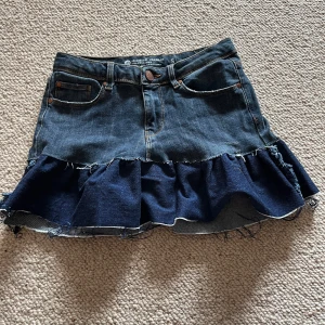 Jeanskjol med volangkant - Mörkblå jeanskjol med volangkant nertill och råa kanter. Klassisk femficksmodell med knapp och dragkedja framtill. Kjolen har en kort längd och är tillverkad i denimtyg.mindre i storleken 