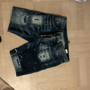 Mörkblå jeansshorts med slitningar och färgstänk - Säljer ett par mörkblå jeansshorts med slitna detaljer och färgstänk i vitt och orange. Shortsen har dragkedja och knapp framtill samt klassiska fickor både fram och bak. Perfekta för en avslappnad och trendig look. Pris kan diskuteras