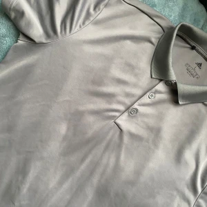 Grå pikétröja från Adidas - Säljer en stilren grå pikétröja från Adidas i återvunnen polyester. Tröjan har klassisk krage, tre knappar och är perfekt för en sportig look. Passar lika bra till träning som till vardags. Adidas-logga på insidan av kragen.