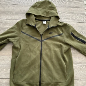 Olivgrön Tröja från Nike - Säljer en olivgrön hoodiejacka från Nike med dragkedja och huva. Jackan har en svart Nike-logga på bröstet, svarta detaljer och en ficka med dragkedja på ärmen. Perfekt för en sportig och avslappnad stil.