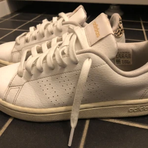 Vita Adidas sneakers med leoparddetalj - Säljer ett par vita Adidas sneakers med snygg leopardmönstrad detalj på hälen och klassisk perforerad sida. Skorna har snörning och platt sula, perfekta för en trendig vardagslook.