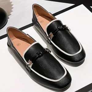 Svarta loafers med vit kant - Snygga svarta loafers med vit kant runt öppningen och dekorativt spänne framtill. Skorna har en klassisk design med rund tå och platt sula, perfekta för en stilren look.