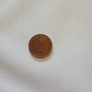 Tysk 10 Pfennig mynt från 1950 - Säljer ett tyskt 10 Pfennig mynt från 1950 med texten 'Bundesrepublik Deutschland' och en ekkvist på ena sidan. Myntet är runt och i kopparfärgat metallmaterial. Perfekt för samlare av äldre europeiska mynt.