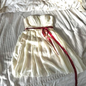 Vit axelbandslös klänning med röd rosett - Säljer en elegant vit axelbandslös klänning med plisserad överdel och röd satinrosett i midjan. Kjolen är lätt och luftig med fina veck. Perfekt för speciella tillfällen. Being / ljus ljus gul.