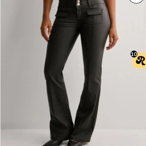 Svarta bootcut byxor i skinnimitation - Snygga svarta byxor med bootcut passform och hög midja. Byxorna har dekorativa fickor framtill med lock och knappar, samt markerade sömmar längs benen. Tillverkade i skinnimitation för en trendig look. Dom är uppsydda för att vara 162-164 cm 