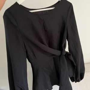 Svart blus - Säljer en svart långärmad blus med rund halsringning och knytband i midjan. Blusen har en enkel och stilren design.