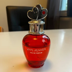 Love Always Eau de Parfum - En parfymflaska i rött glas med silverfärgat lock dekorerat med två hjärtan, varav ett har en röd detalj. Flaskan har texten 'Love Always Eau de Parfum' tryckt i vitt på framsidan. Doften är romantisk och flaskan har en rund, elegant form.