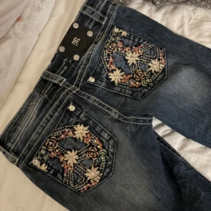 Miss Me jeans med broderade bakfickor - Säljer ett par mörkblå Miss Me jeans med snygga broderade detaljer och stenar på bakfickorna. Byxorna har låg midja och bootcut-modell, perfekt för dig som gillar unika detaljer och en klassisk jeanslook.