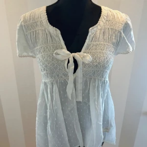 Vit blus med knyt från Odd Molly - Supersöt vit blus från Odd Molly med korta ärmar, smockdetaljer och knyt framtill. Blusen har diskret prickigt mönster i tyget och en luftig, romantisk känsla. Perfekt till sommaren och vårens alla tillfällen! Säljer för att den är för stor på mig
