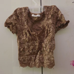 Brun spetsblus - Säljer en charmig brun spetsblus med korta ärmar. Blusen har ett vackert blommigt mönster och en elegant design som passar perfekt för en stilfull look. Perfekt för att lägga till en touch av elegans i din garderob.