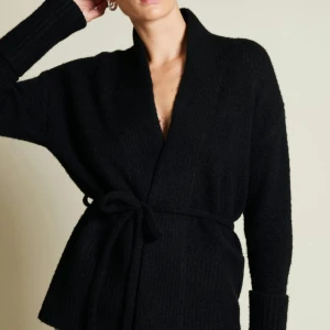 Svart stickad kofta med knytband  - Säljer en svart stickad kofta med knytband i midjan. Koftan har långa ärmar och en mjuk, bekväm passform. 