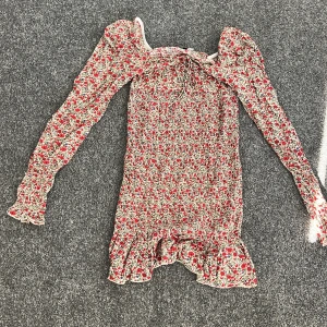 Blommig klänning från NA-KD - Säljer en söt blommig klänning från NA-KD i storlek 36. Klänningen har långa ärmar och en volangdetalj vid fållen. Perfekt för vår och sommar med sitt färgglada mönster i rött och blått på en beige bakgrund.
