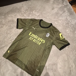 Grön fotbollströja från Puma - Säljer en grön fotbollströja från Puma med korta ärmar. Tröjan har svarta detaljer och sponsortexten 'Emirates Fly Better' samt 'BitMEX' tryckt i gult. Perfekt för fotbollsfans som vill ha en stilren och modern look.