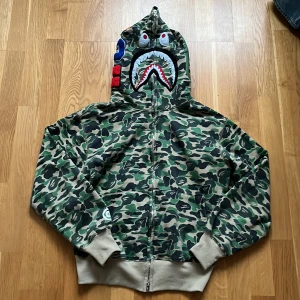 Camouflage hoodie från A Bathing Ape - Säljer en cool camouflage hoodie från A Bathing Ape med hajmotiv på huvan. Den har dragkedja och ribbade muddar. Perfekt för streetwear-stilen! Passar S också och är Riktig Bape!