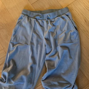 Blå mjukisbyxor med fickor - Säljer ett par bekväma blå mjukisbyxor från Juicy med elastisk midja och snörning. Byxorna har praktiska fickor fram och bak, samt dekorativa knappar på bakfickorna. Perfekta för en avslappnad stil.