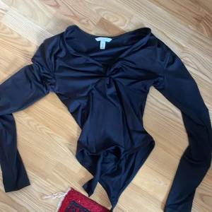 Svart långärmad topp - Snygg svart långärmad body med en unik twistdetalj framtill. Tillverkad i ett glansigt material som ger en elegant look. Perfekt för en kväll ute eller en speciell tillställning.