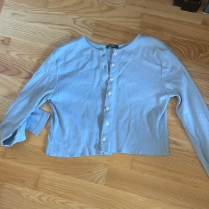 Ljusblå kofta med knappar - Säljer en ljusblå kofta med knappar framtill. Den har en enkel och stilren design med långa ärmar. Perfekt för lager-på-lager-stil eller som ett lättare ytterplagg.