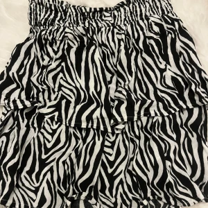 Zebramönstrad kortkjol - Snygg kortkjol med zebramönster i svart och vitt. Kjolen har en elastisk midja och volanger som ger en lekfull look. Perfekt för att sticka ut i mängden!