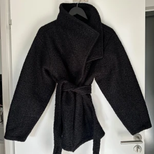 Svart kappa från Gina Tricot - Snygg svart kappa från Gina Tricot med knytbälte i midjan. Den har en stilren design med långa ärmar och en mjuk, ullig textur. Perfekt för vårdagar när du vill hålla stilen. Endast använd en gång! 🖤