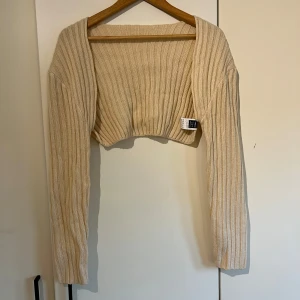Beige ribbad bolerotopp från SHEIN - Säljer en beige ribbad bolerotopp från SHEIN. Den är croppad och har långa ärmar, perfekt för att lägga till en stilren touch till din outfit. Tillverkad i 100% akryl, vilket gör den både mjuk och bekväm. Passar bra till både vardag och speciella tillfällen.