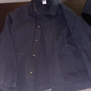 Svart  jacka från Nike - Säljer en svart jacka från Nike med knappar framtill och två fickor. Jackan har en klassisk krage och är perfekt för en avslappnad stil. Passar bra till både vardag och träning.