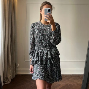 Blommig klänning från Isabel Étoile - Säljer en söt blommig klänning från Isabel Étoile med volanger och långa ärmar. Klänningen har en mörk bas med ljusa blommor i rosa och blått. Perfekt för en stilfull look!