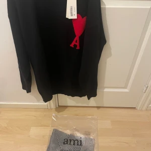 Svart tröja med röd detalj från Ami - Säljer en svart tröja från Ami med en stor röd hjärtformad detalj och bokstaven 'A' på bröstet. Tröjan har långa ärmar och en klassisk passform. Perfekt för en stilren look.
